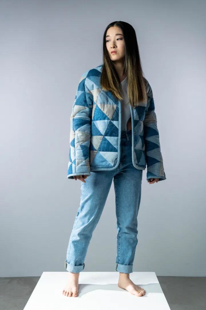 Model berdiri di atas kotak putih memakai jaket tambal segitiga dan jeans biru muda yang digulung