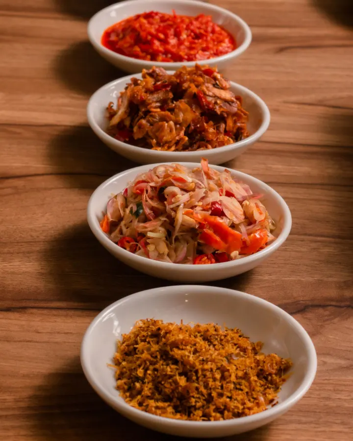 Five white bowls of sambal in a row on a wooden table, from dark roasted chilli to shallot sambal and beef floss